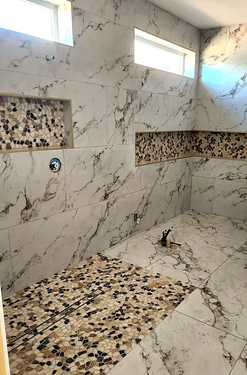 Marble-look bathroom with a pebble mosaic accent band through the niche and a matching strip at the linear drain